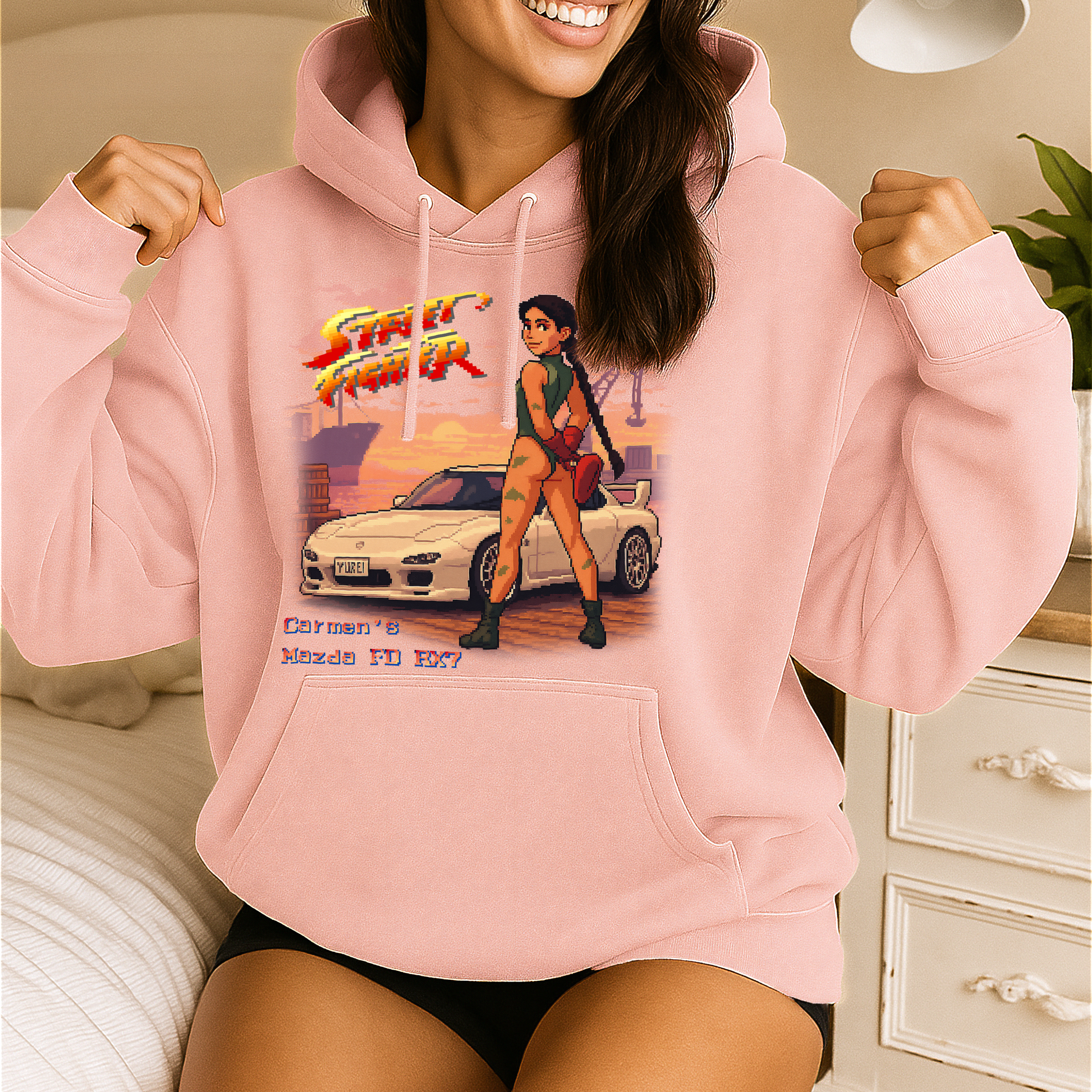 A smiling woman poses indoors wearing a pastel pink hoodie featuring a custom Street Fighter II–inspired pixel art design. The graphic showcases a white Mazda FD RX-7 with the number plate “YUREI” and a character modeled after Cammy standing confidently in front of the car at sunset. Text below reads “Carmen’s Mazda FD RX7,” blending retro gaming aesthetics with car culture.