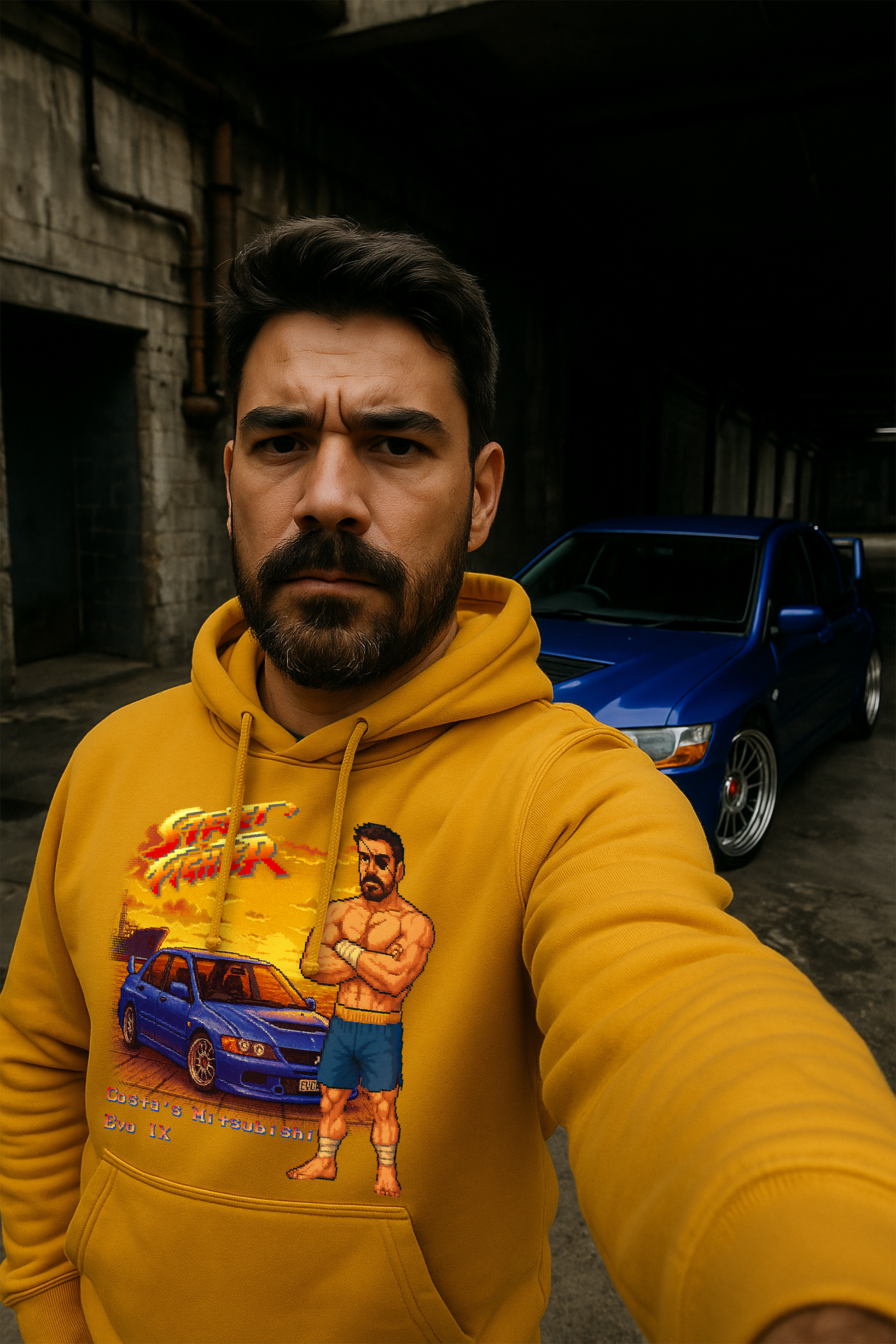 A man in a gold hoodie takes a selfie in front of his blue Mitsubishi Lancer Evolution IX, parked in a gritty industrial setting. The hoodie features a bold pixel-art Street Fighter II–inspired design of the same car with a custom character modeled after the wearer, styled like Sagat—topless, muscular, with an eyepatch and Muay Thai shorts—standing in front of a sunset dock scene.