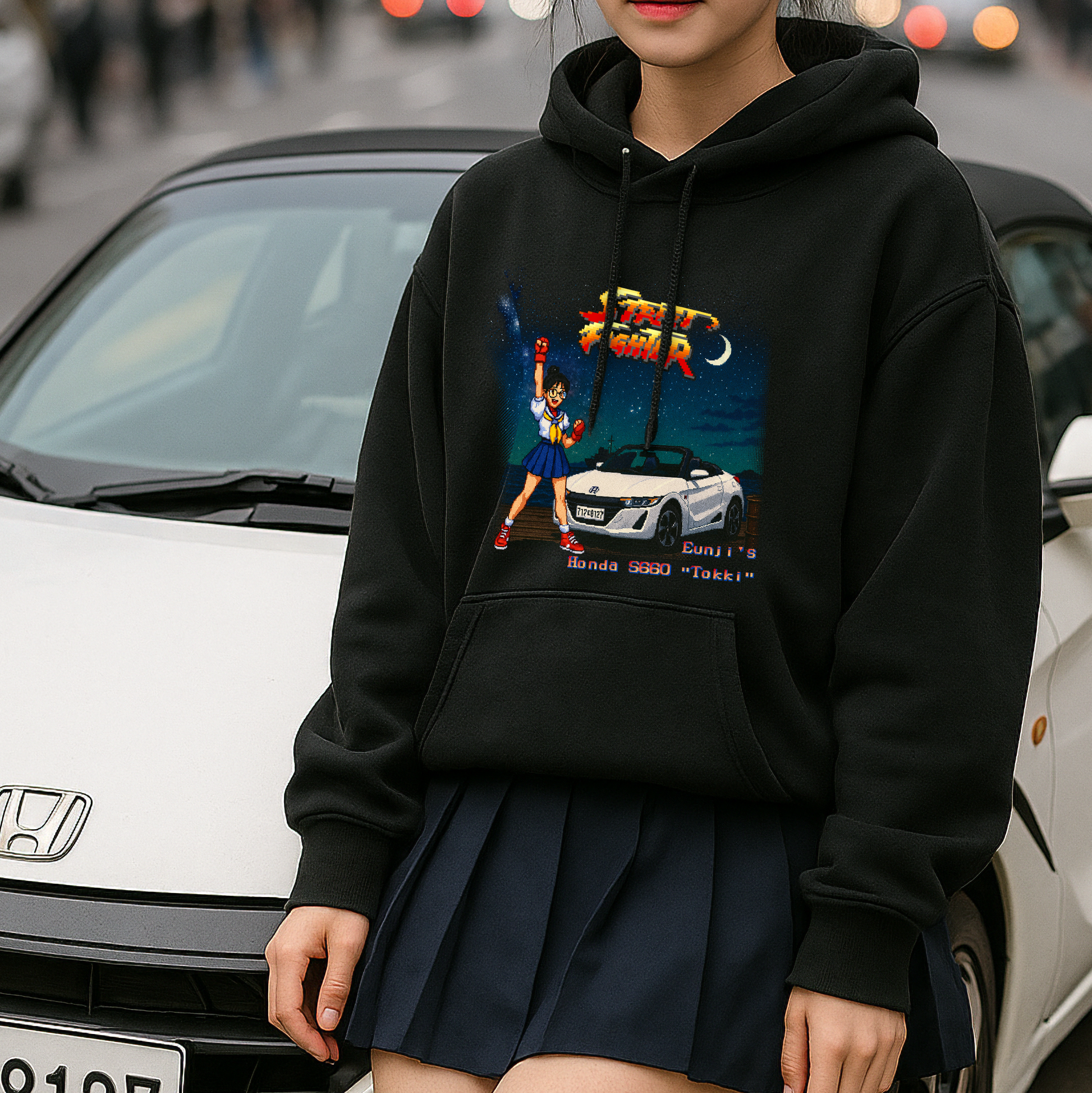 Young woman in black hoodie and pleated skirt stands in front of a white Honda S660 convertible on a city street. Her hoodie features a retro 16-bit pixel art graphic inspired by the Street Fighter II bonus stage, showing a Sakura-style fighter next to the car under a twilight sky. A unique blend of Japanese car culture and classic arcade game nostalgia.

Let me know if you'd like a shorter or SEO-optimized variant for Shopify or Etsy!