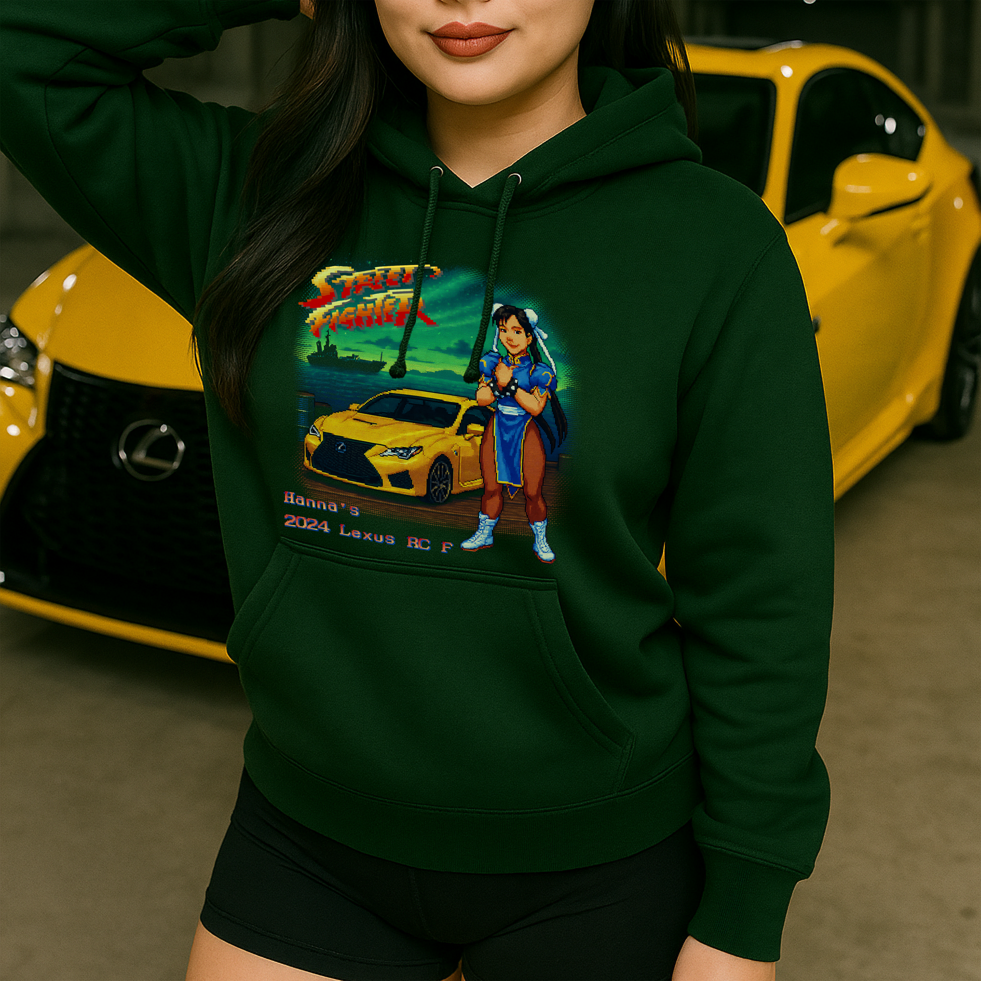 Young woman wearing a forest green hoodie featuring a retro 16-bit pixel art scene inspired by Street Fighter II. The design showcases Chun-Li standing proudly beside a bright yellow 2024 Lexus RC F on a waterfront dock at dusk. The real-life car in the background matches the graphic, blending gaming nostalgia with modern car culture.