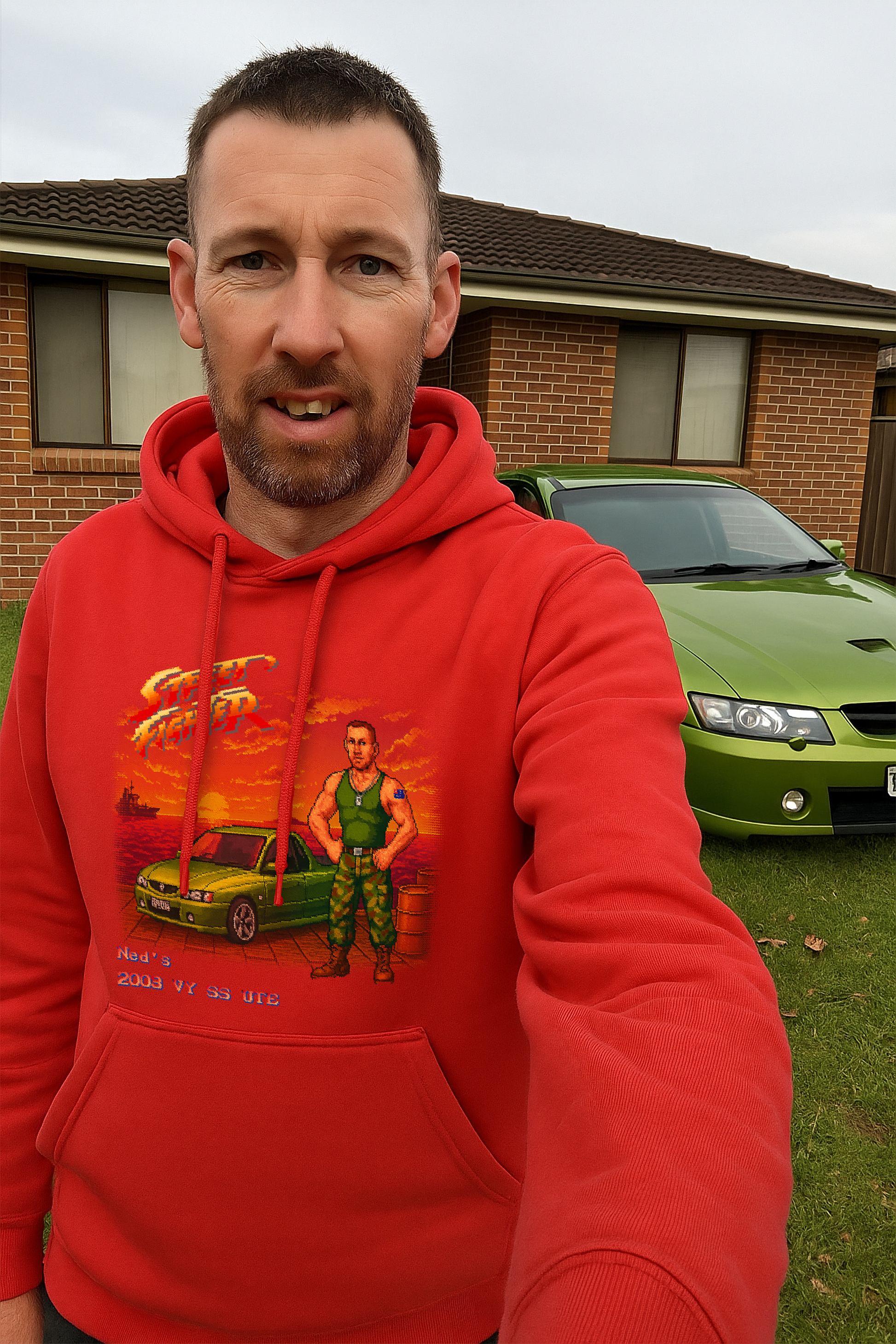A man takes a selfie in front of his lime green 2003 Holden VY SS Ute, parked on a grassy lawn outside a brick home in suburban Australia. He wears a red hoodie featuring a Street Fighter II–style pixel art design with a custom character inspired by Guile—complete with dog tags, camo pants, combat boots, and an Australian flag tattoo—standing next to the same green ute on a dock at sunset. The text on the hoodie reads “Ned’s 2003 VY SS UTE.” The image blends retro gaming and Aussie car culture