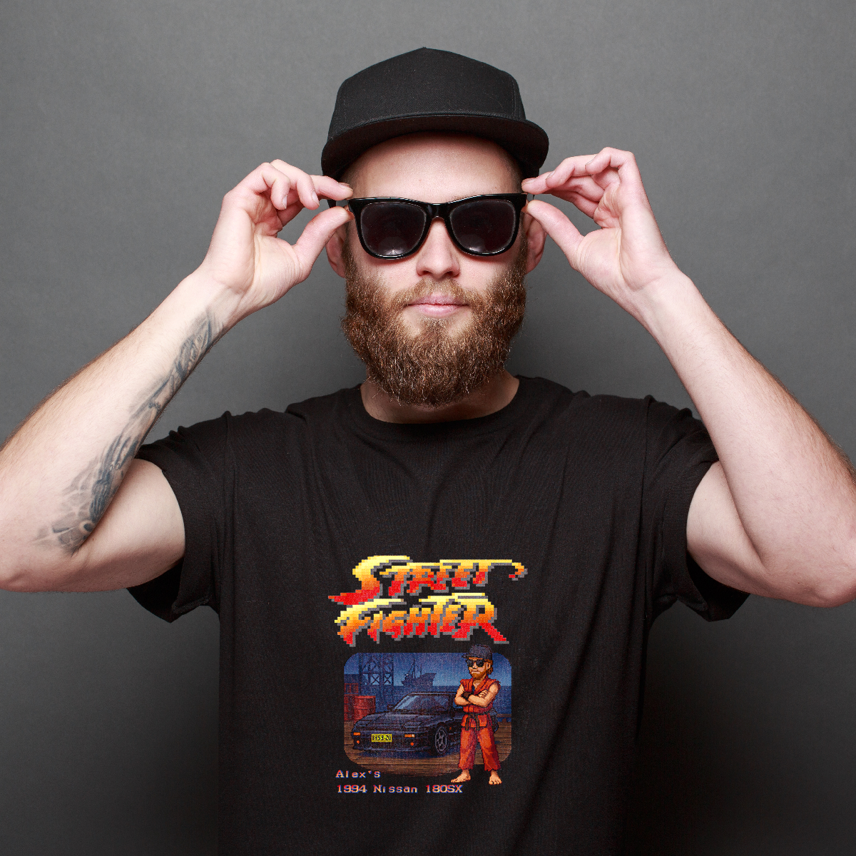 Bearded man wearing cap and sunglasses models a custom black t-shirt featuring personalized pixel art of himself in a red gi beside his 1994 Nissan 180SX, styled after a retro fighting game.
