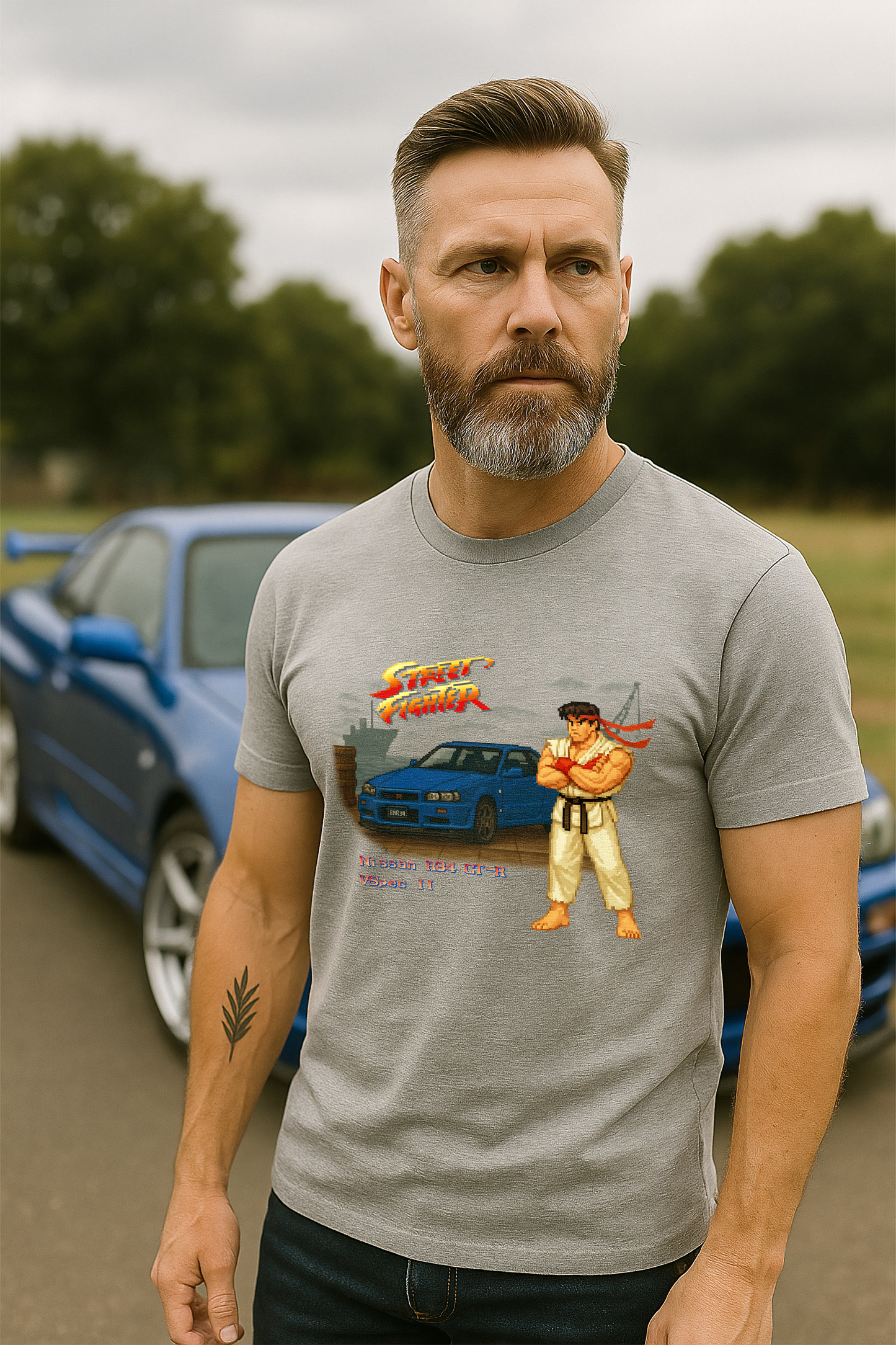 A fit, middle-aged man with a salt-and-pepper beard wears a grey marle t-shirt while standing in front of a Bayside Blue Nissan R34 GT-R V-Spec II on an overcast day. The t-shirt features a retro Street Fighter II–inspired pixel art design with Ryu standing beside the R34 on a dock background, complete with the Street Fighter logo and text that reads “Nissan R34 GT-R Vspec II.” The scene combines 90s arcade nostalgia with JDM car culture.