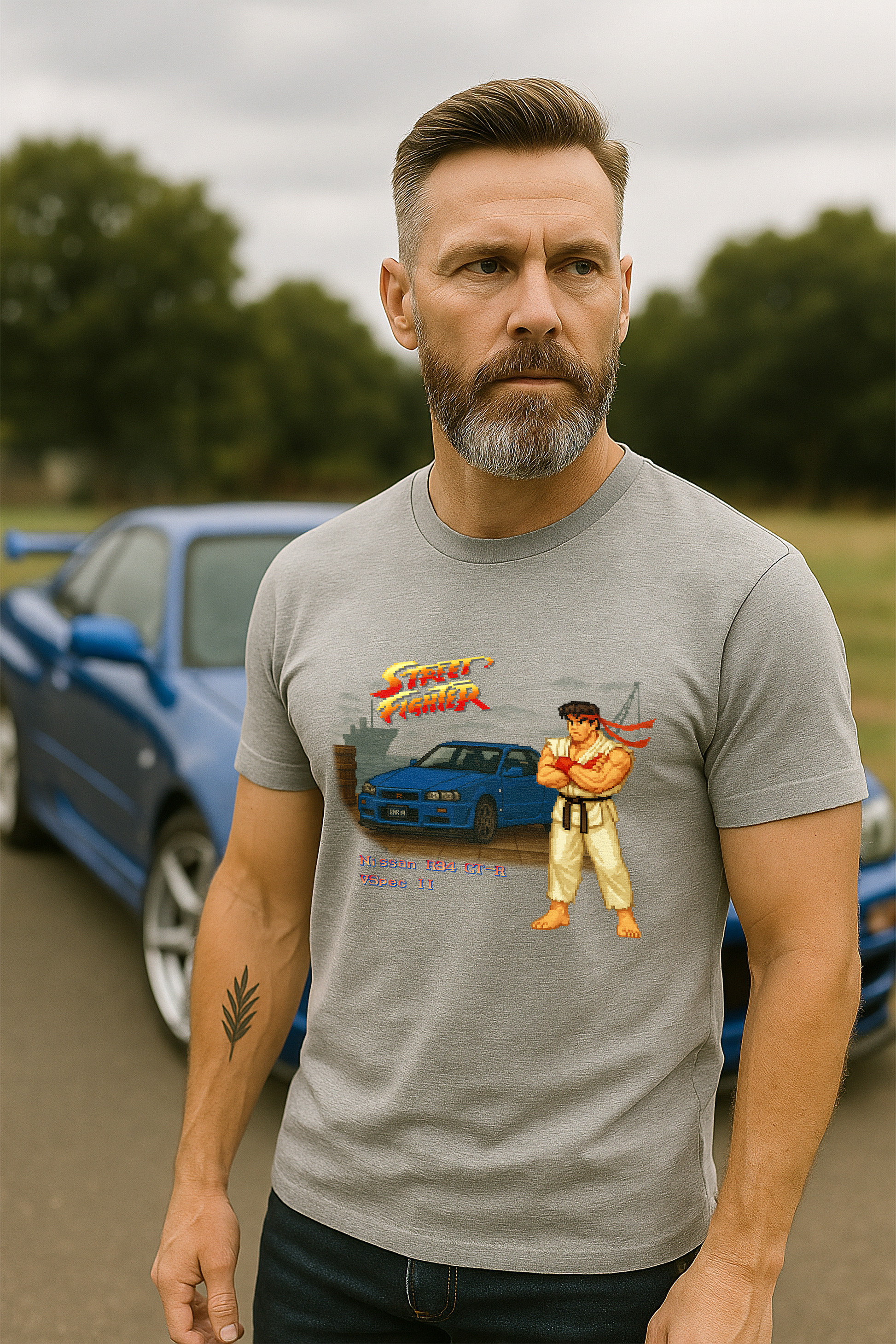 A fit, middle-aged man with a salt-and-pepper beard wears a grey marle t-shirt while standing in front of a Bayside Blue Nissan R34 GT-R V-Spec II on an overcast day. The t-shirt features a retro Street Fighter II–inspired pixel art design with Ryu standing beside the R34 on a dock background, complete with the Street Fighter logo and text that reads “Nissan R34 GT-R Vspec II.” The scene combines 90s arcade nostalgia with JDM car culture.