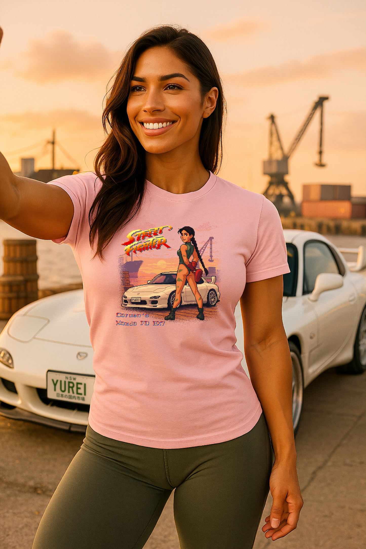 Woman wearing a pink Street Fighter II pixel art T-shirt featuring custom 16-bit design of a white Mazda RX-7 FD and Cammy-inspired character. Text reads "Carmen’s Mazda FD RX7." Shot at golden hour beside the real RX-7 with “YUREI” license plate. Ideal for JDM lovers, retro arcade fans, and unique anime gamer gift ideas.