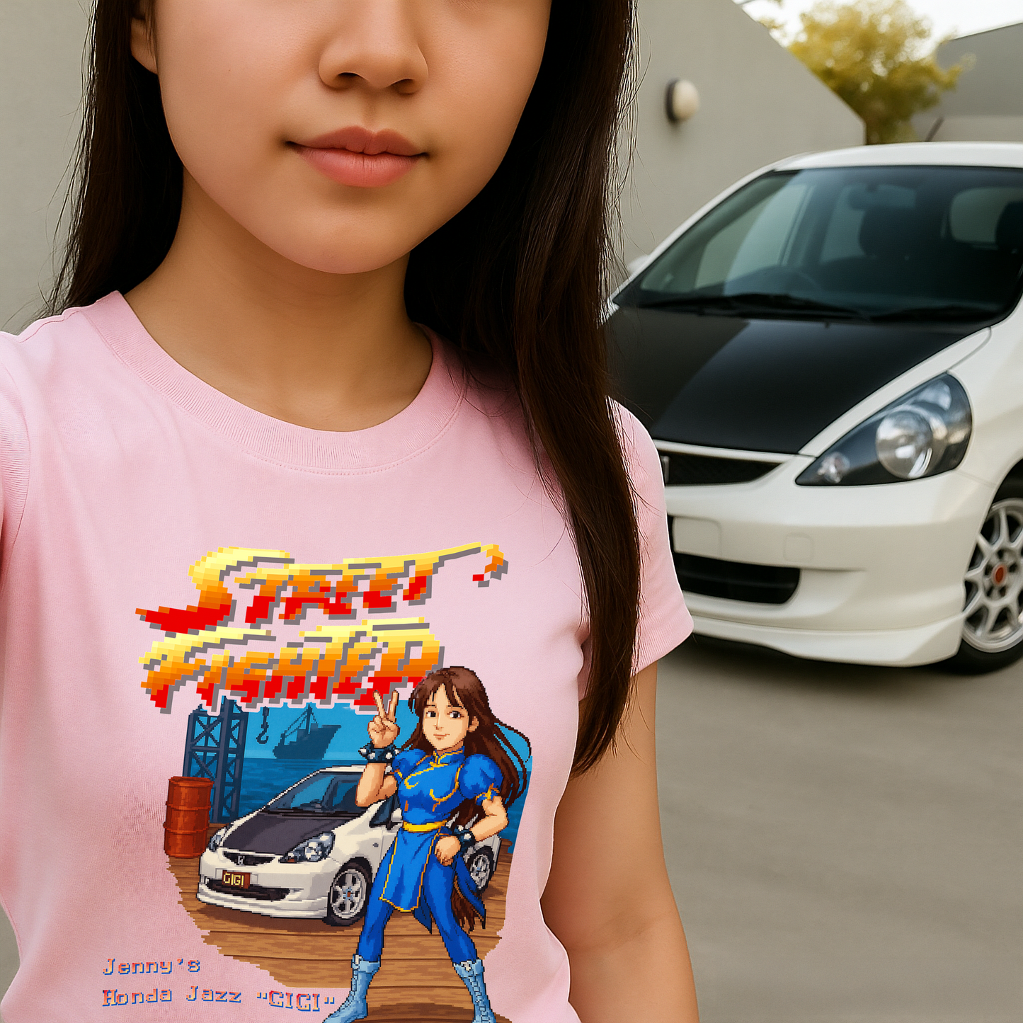 Pink Street Fighter II pixel art T-shirt featuring custom 16-bit design of a white Honda Jazz and Chun-Li-inspired character. Text reads "Jenny’s Honda Jazz 'GIGI'." Worn by young woman posing in front of the real white Honda Jazz. Ideal for retro arcade fans, JDM enthusiasts, and personalized gamer gifts.