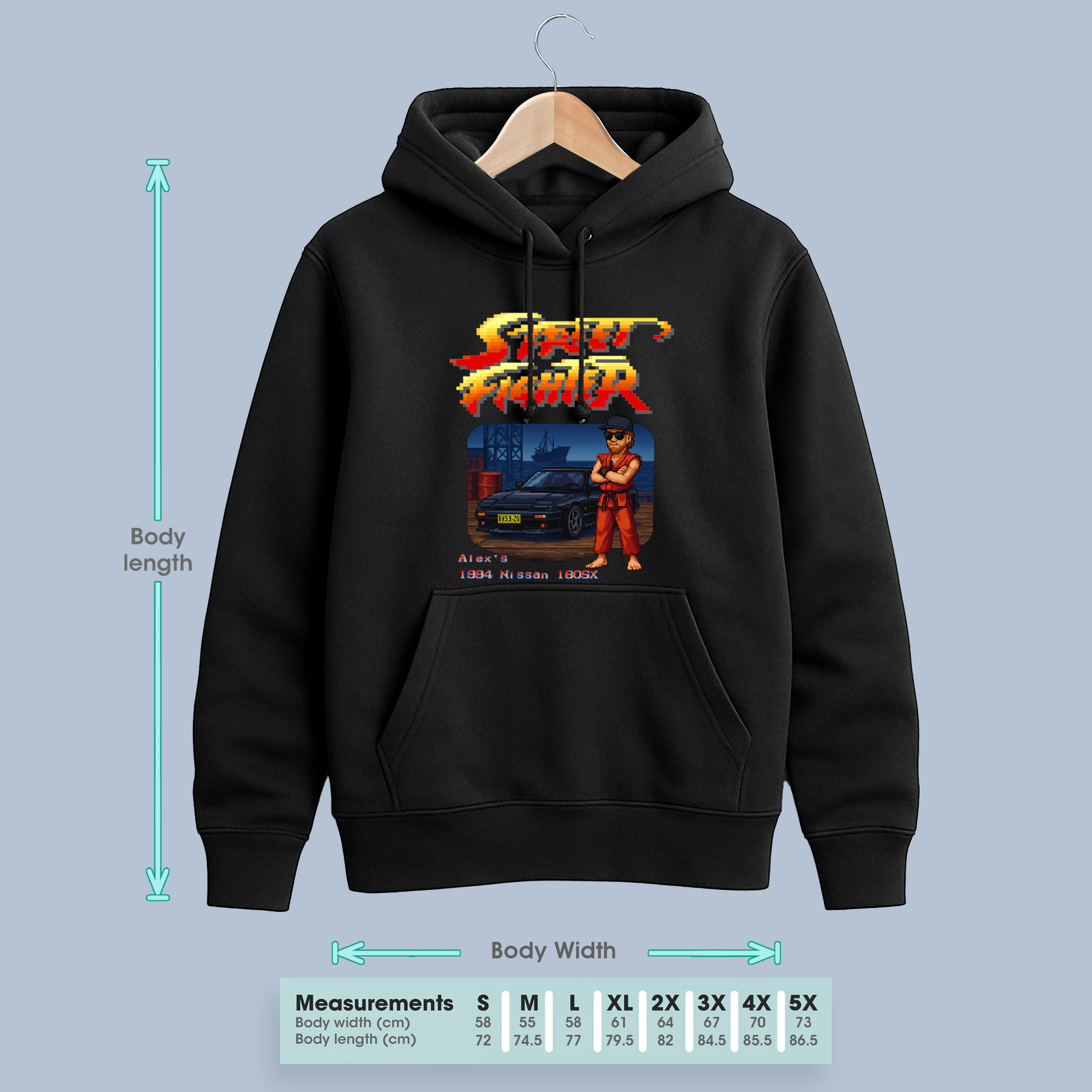 Black hoodie with custom Street Fighter-style pixel art hanging on a coathanger against a blank wall, displayed next to a size chart for measurements.